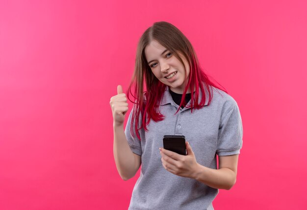 Mujer hermosa joven alegre que lleva la camiseta gris que sostiene el teléfono que muestra el gesto sí en la pared rosada aislada