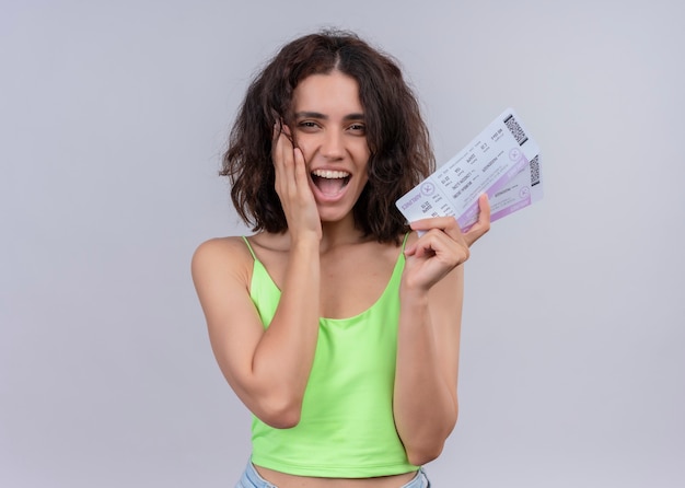 Mujer hermosa joven alegre que sostiene los billetes de avión y que pone la mano en la mejilla en la pared blanca aislada