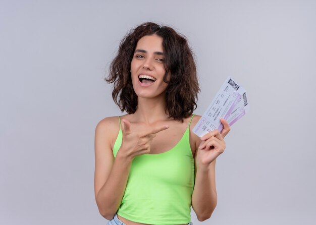 Mujer hermosa joven alegre que sostiene los billetes de avión y que los señala en la pared blanca aislada