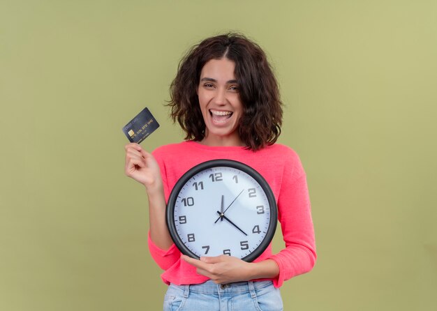 Mujer hermosa joven alegre que sostiene la tarjeta y el reloj en la pared verde aislada