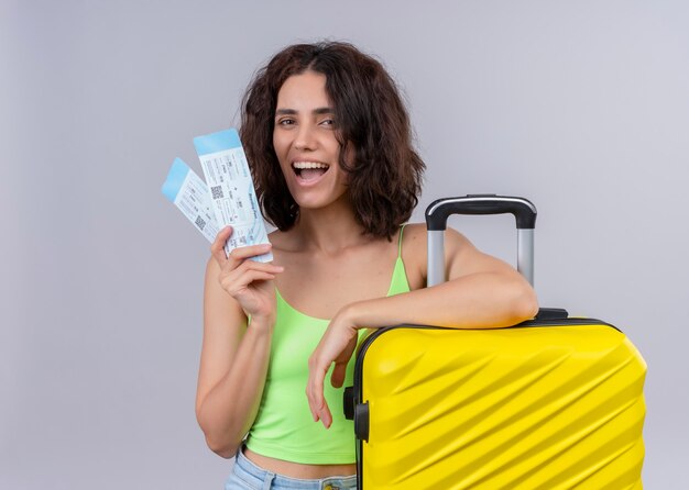 Mujer hermosa joven alegre del viajero que sostiene los billetes de avión y la maleta en la pared blanca aislada