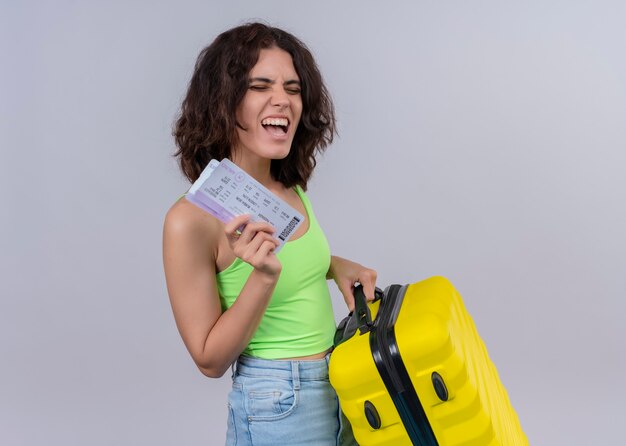 Mujer hermosa joven alegre del viajero que sostiene los billetes de avión y la maleta en la pared blanca aislada