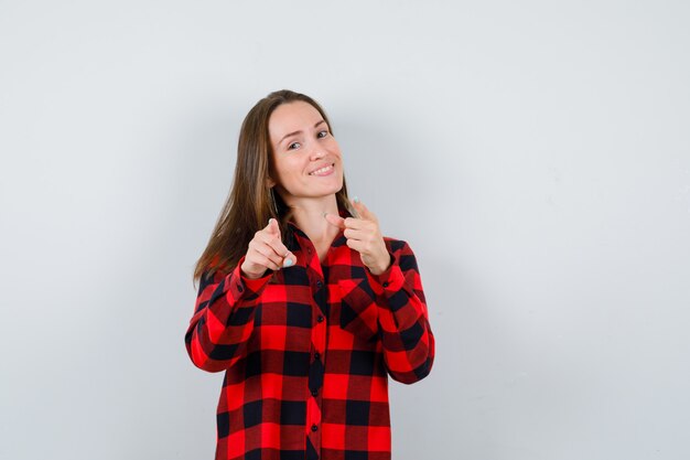 Mujer hermosa joven apuntando al frente en camisa casual y mirando alegre. vista frontal.