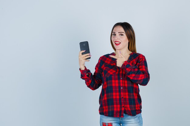 Mujer hermosa joven apuntando al teléfono en camisa casual y mirando dichoso. vista frontal.