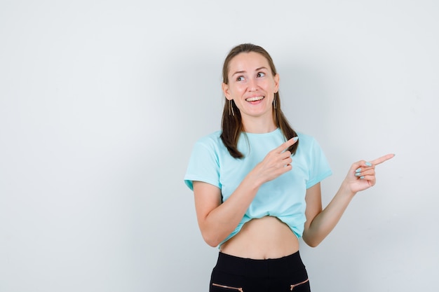 Mujer hermosa joven apuntando a la esquina superior derecha, mirando a otro lado en camiseta y mirando feliz, vista frontal.