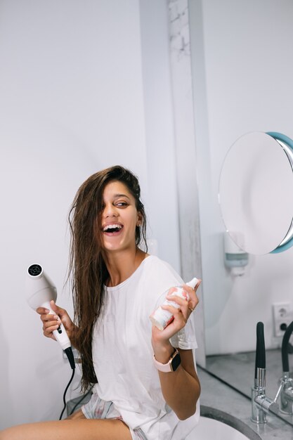 Mujer hermosa joven en el baño sosteniendo un secador de pelo y una botella pequeña, vista cercana