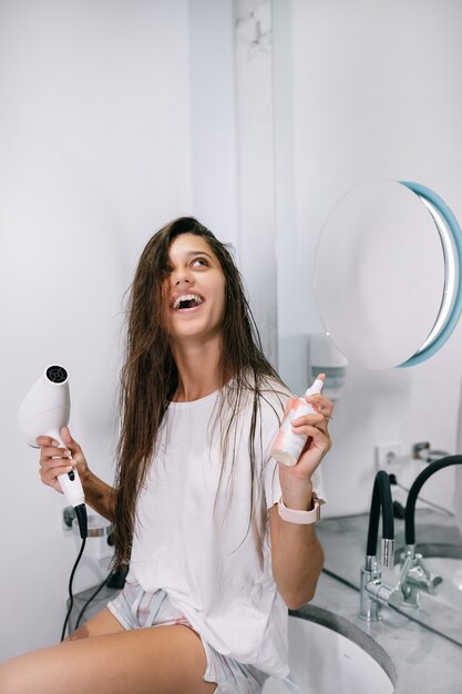 Mujer hermosa joven en el baño sosteniendo un secador de pelo y una botella pequeña