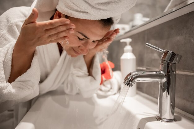 Mujer hermosa joven en el baño con una toalla en la cabeza se lava la cara con agua del grifo