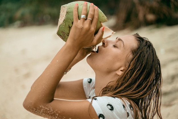 Mujer hermosa joven bebe agua de coco de coco en la playa.