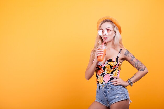 Mujer hermosa joven bebiendo jugo de naranja con sombrero y gafas de sol en estudio