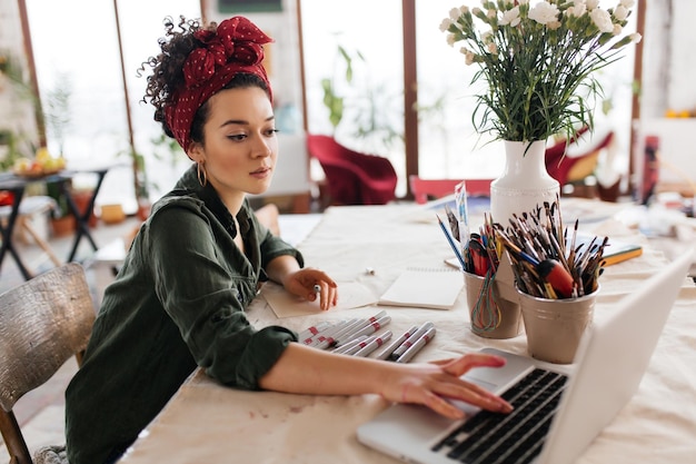 Mujer hermosa joven con cabello rizado oscuro sentada en la mesa soñadoramente usando una computadora portátil y dibujando bocetos pasando tiempo en un taller moderno y acogedor con grandes ventanas