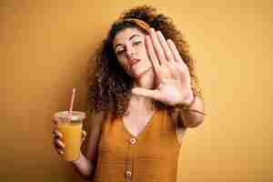 Foto gratuita mujer hermosa joven con cabello rizado y piercing bebiendo jugo de naranja saludable con la mano abierta haciendo señas de alto con gesto de defensa de expresión seria y segura