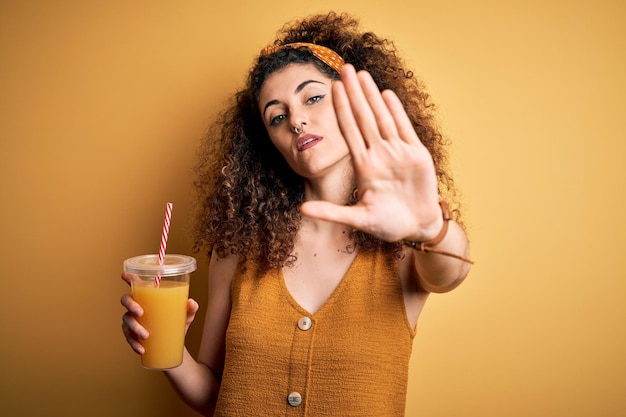 Mujer hermosa joven con cabello rizado y piercing bebiendo jugo de naranja saludable con la mano abierta haciendo señas de alto con gesto de defensa de expresión seria y segura