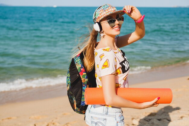 Mujer hermosa joven caminando en la playa con estera de yoga, escuchando música en auriculares, estilo swag deportivo hipster, pantalones cortos de mezclilla, camiseta, mochila, gorra, gafas de sol, soleado, fin de semana de verano, alegre