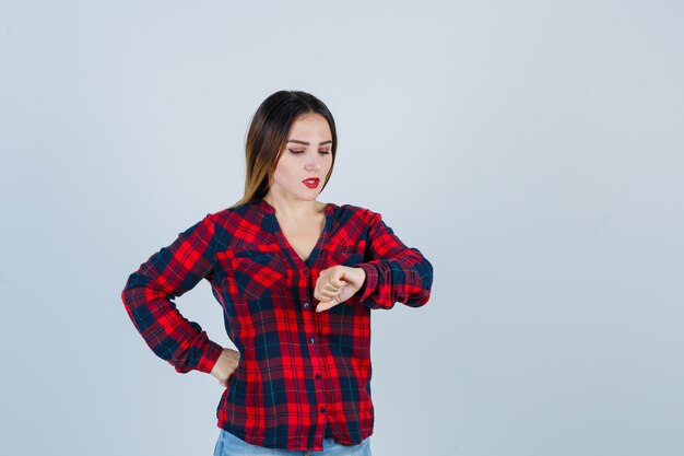 Mujer hermosa joven en camisa casual fingiendo mirar el reloj en su muñeca y mirando enfocado, vista frontal.