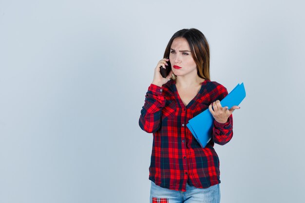 Mujer hermosa joven en camisa casual, jeans sosteniendo la carpeta mientras habla por teléfono, apuntando al frente y mirando perplejo, vista frontal.