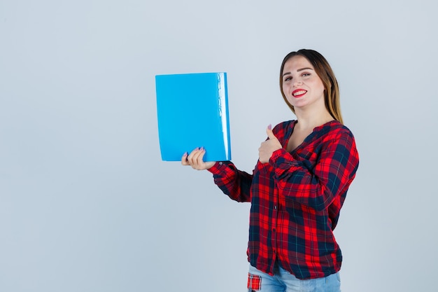 Mujer hermosa joven en camisa casual, jeans sosteniendo la carpeta, mostrando el pulgar hacia arriba y mirando alegre, vista frontal.