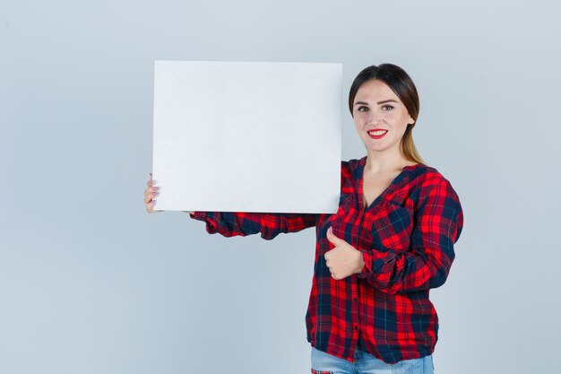 Mujer hermosa joven en camisa casual con tablero en blanco, mostrando el pulgar hacia arriba y mirando positivo, vista frontal.