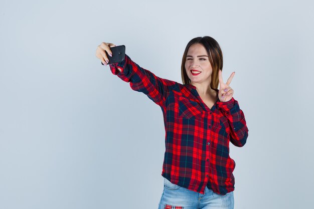 Mujer hermosa joven en camisa casual tomando selfie, mostrando gesto de paz y mirando alegre, vista frontal.