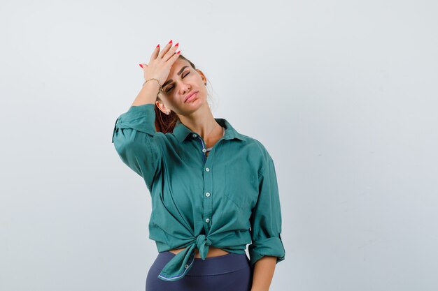 Mujer hermosa joven en camisa verde manteniendo la mano en la frente y mirando soñoliento, vista frontal.