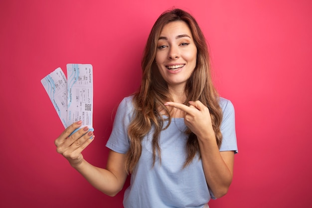 Mujer hermosa joven en camiseta azul con billetes de avión apuntando con el dedo índice a ellos sonriendo alegremente