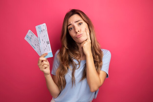 Mujer hermosa joven en camiseta azul con billetes de avión mirando a la cámara confundida y disgustada de pie sobre fondo rosa