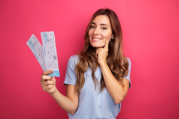 Mujer hermosa joven en camiseta azul con billetes de avión mirando a la cámara sonriendo alegremente de pie sobre fondo rosa