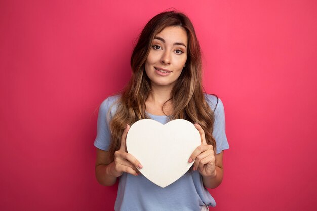 Mujer hermosa joven en camiseta azul con corazón de cartón mirando a la cámara sonriendo alegremente de pie sobre fondo rosa