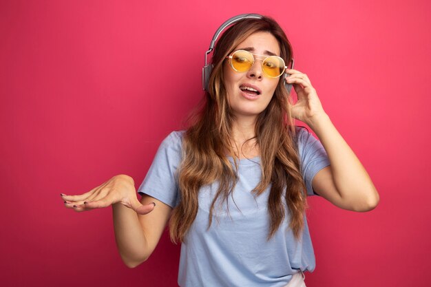 Mujer hermosa joven en camiseta azul con gafas amarillas con auriculares mirando a la cámara gesticulando con las manos de pie autosatisfecho sobre fondo rosa