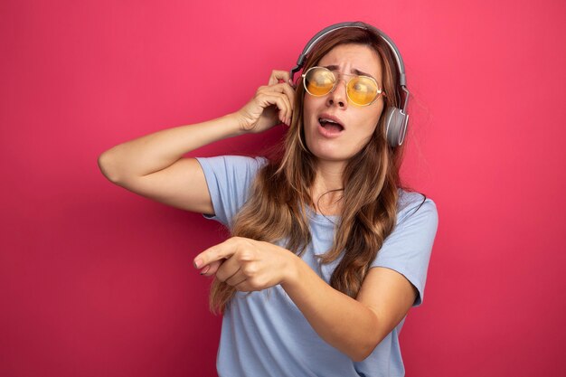 Mujer hermosa joven en camiseta azul con gafas amarillas con auriculares mirando a un lado confundido apuntando con el dedo índice hacia el lado