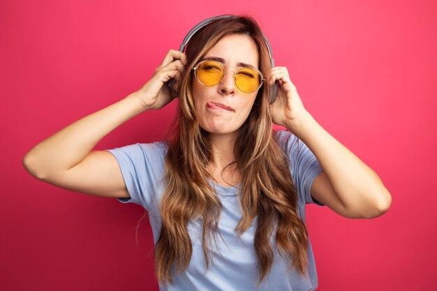 Mujer hermosa joven en camiseta azul con gafas amarillas con auriculares mirando a un lado feliz y alegre sacando la lengua de pie sobre fondo rosa