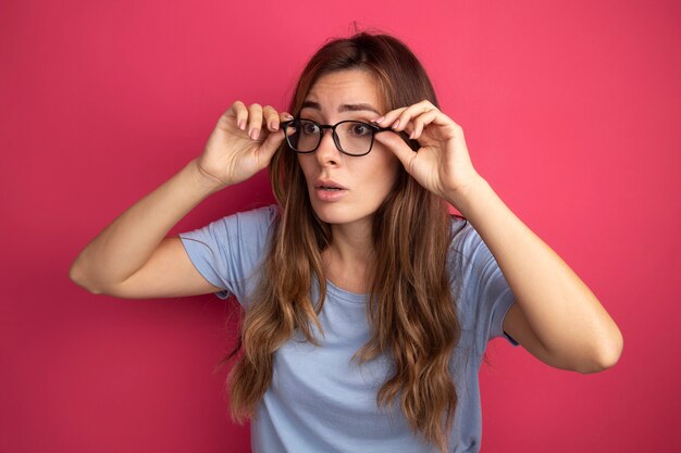 Mujer hermosa joven en camiseta azul con gafas mirando a un lado de cerca siendo sorprendido de pie sobre fondo de color rosa