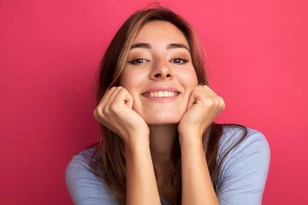 Mujer hermosa joven en camiseta azul mirando a la cámara con la cabeza apoyada en los puños feliz y positivo sonriendo de pie sobre rosa