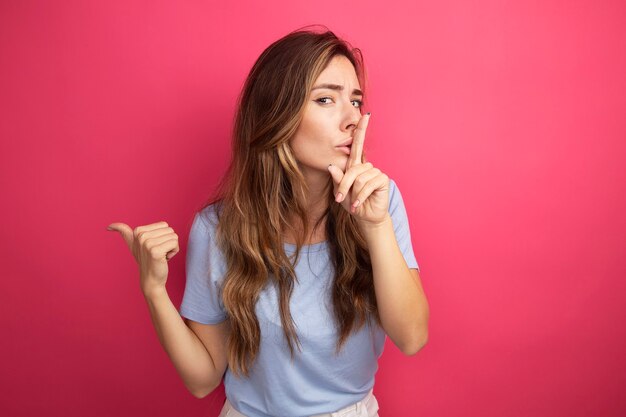 Mujer hermosa joven en camiseta azul mirando a la cámara haciendo gesto de silencio con el dedo en los labios apuntando con el pulgar hacia el lado