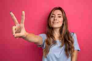 Foto gratuita mujer hermosa joven en camiseta azul mirando a un lado con una sonrisa en la cara mostrando v-sign de pie sobre fondo rosa