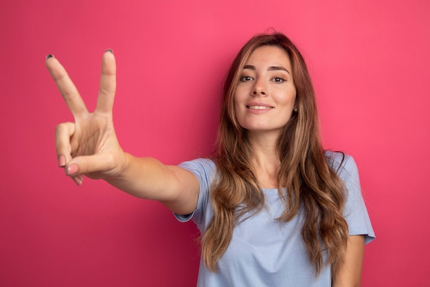 Mujer hermosa joven en camiseta azul mirando a un lado con una sonrisa en la cara mostrando v-sign de pie sobre fondo rosa