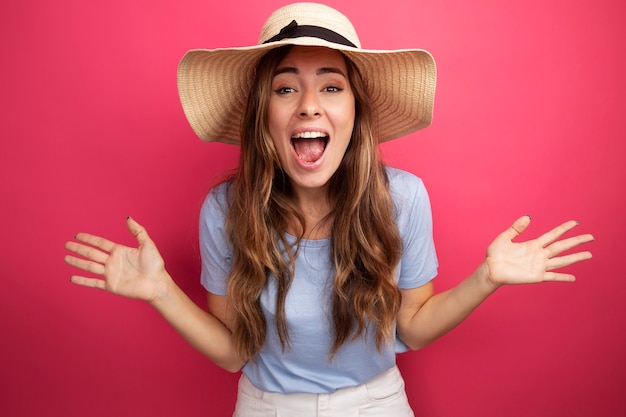 Mujer hermosa joven en camiseta azul y sombrero de verano mirando a la cámara loco feliz y emocionado gritando levantando los brazos sobre fondo rosa