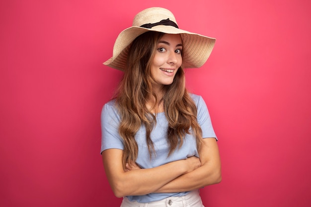 Mujer hermosa joven en camiseta azul y sombrero de verano mirando a la cámara sonriendo feliz y positivo con los brazos cruzados