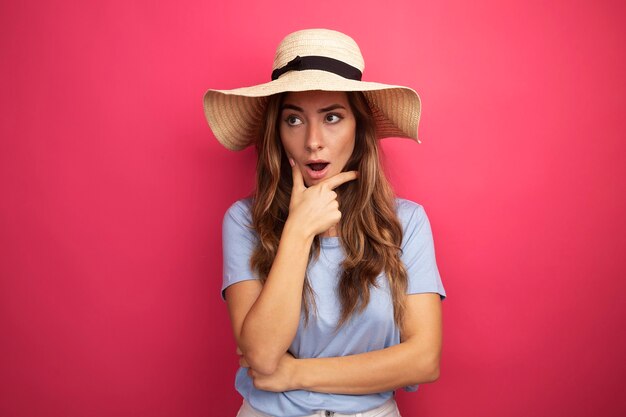 Mujer hermosa joven en camiseta azul y sombrero de verano mirando a un lado sorprendido de pie sobre fondo rosa