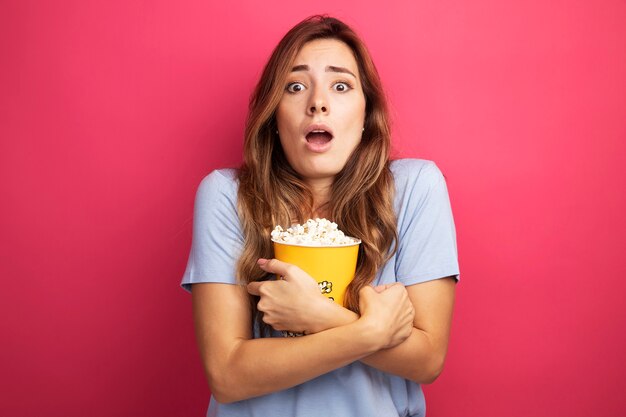 Mujer hermosa joven en camiseta azul sosteniendo un cubo con palomitas de maíz mirando a la cámara sorprendida de pie sobre fondo rosa