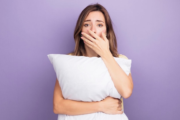 Foto gratuita mujer hermosa joven en camiseta beige con almohada blanca mirando a la cámara conmocionado cubriendo la boca con la mano