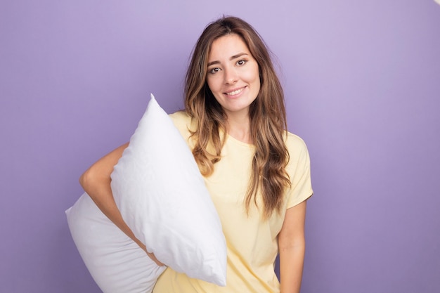 Mujer hermosa joven en camiseta beige con almohada blanca mirando a la cámara con una sonrisa en la cara de pie sobre púrpura