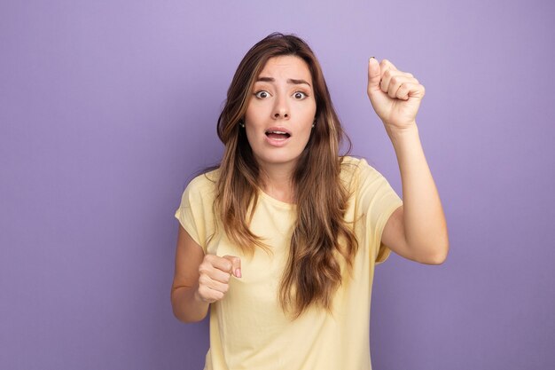 Mujer hermosa joven en camiseta beige mirando a cámara confundida levantando puños sobre púrpura