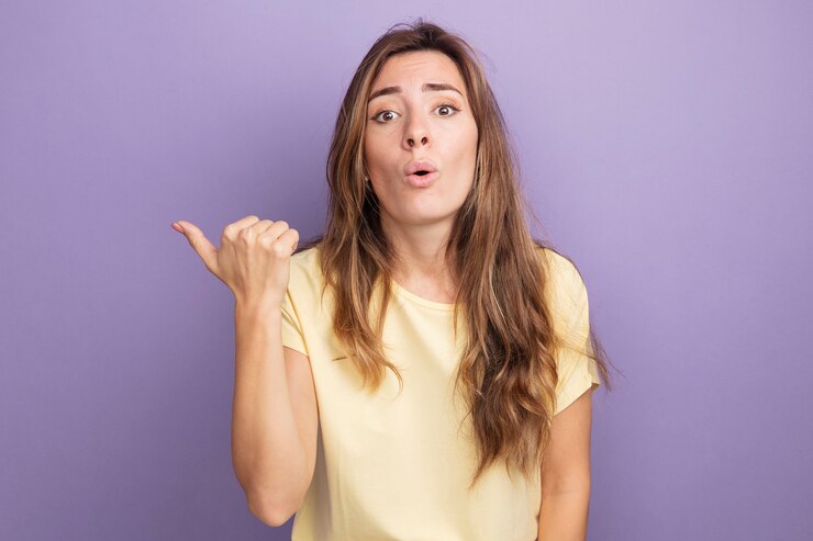 Foto gratuito mujer hermosa joven en camiseta beige mirando a cámara confundida y sorprendida apuntando con el pulgar hacia atrás