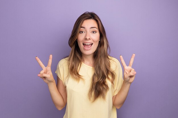 Mujer hermosa joven en camiseta beige mirando a cámara feliz y emocionado mostrando v-sign de pie sobre púrpura