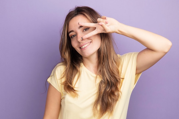 Mujer hermosa joven en camiseta beige mirando a la cámara sonriendo feliz y alegre mostrando v-sign cerca de su ojo de pie sobre fondo púrpura