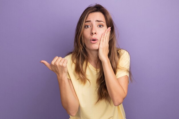 Mujer hermosa joven en camiseta beige mirando a cámara sorprendida y confundida con la mano en la mejilla apuntando con el pulgar hacia el lado de pie sobre fondo púrpura