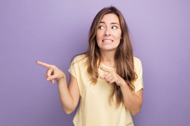 Mujer hermosa joven en camiseta beige mirando a un lado confundido apuntando con figuras índice al lado de pie sobre púrpura