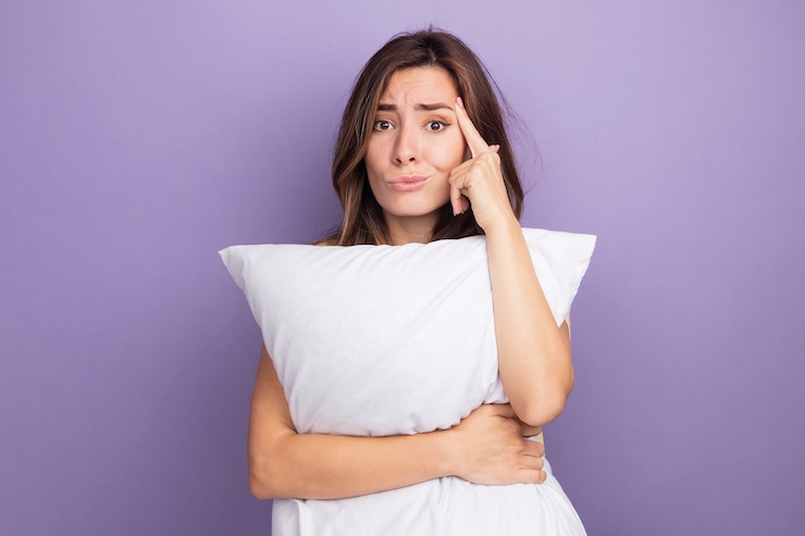 Foto gratuito mujer hermosa joven en camiseta beige sosteniendo una almohada blanca mirando a la cámara confundida y disgustada apuntando a su sien