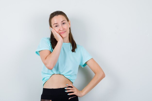 Mujer hermosa joven en camiseta con la mano en la cara mientras posa y mira alegre, vista frontal.